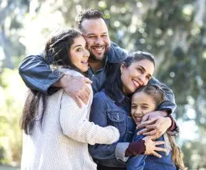 An Hispanic family of four, with two girls, standing outdoors at the park on a sunny autumn day, smiling, looking away from the camera and hugging one another. The younger daughter, 9 years old, is standing next to the mother. Her sister, a 13 year old teenager, is standing next to father.