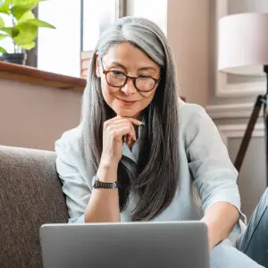 Older woman with long straight grey hair and glasses sitting on her couch and smiling while looking at her laptop