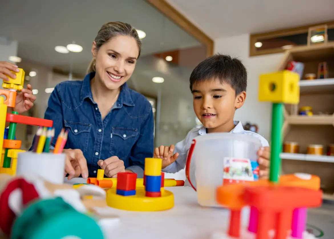 Happy Latin American schoolboy playing with building blocks in the classroom and the supervision of his teacher