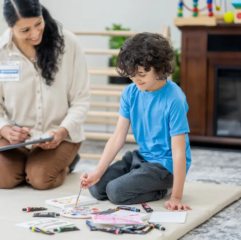 A young boy lays out on the floor with various art supplies in front of him as he works with an Occupational Therapist. The Therapist is seated beside him and is holding a tablet as she takes notes during the appointment.