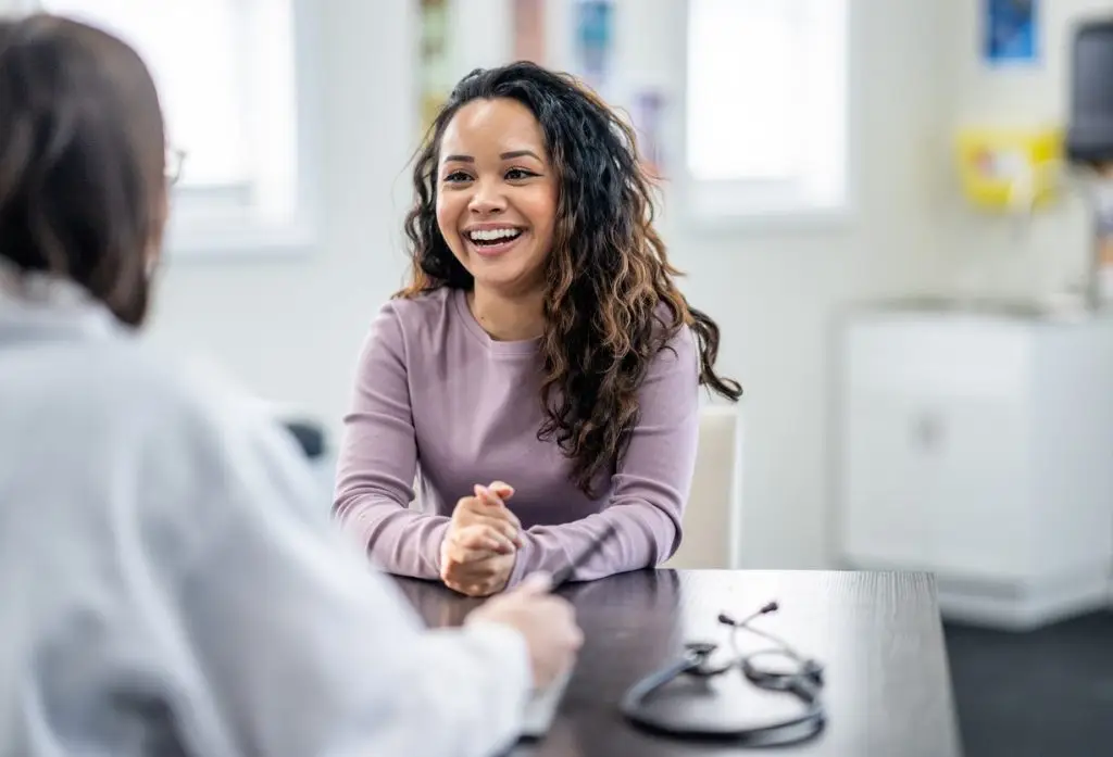 A young woman of mixed race, sits with her female doctor as they discuss her health concerns. The patient is dressed casually and has a smile on her face.