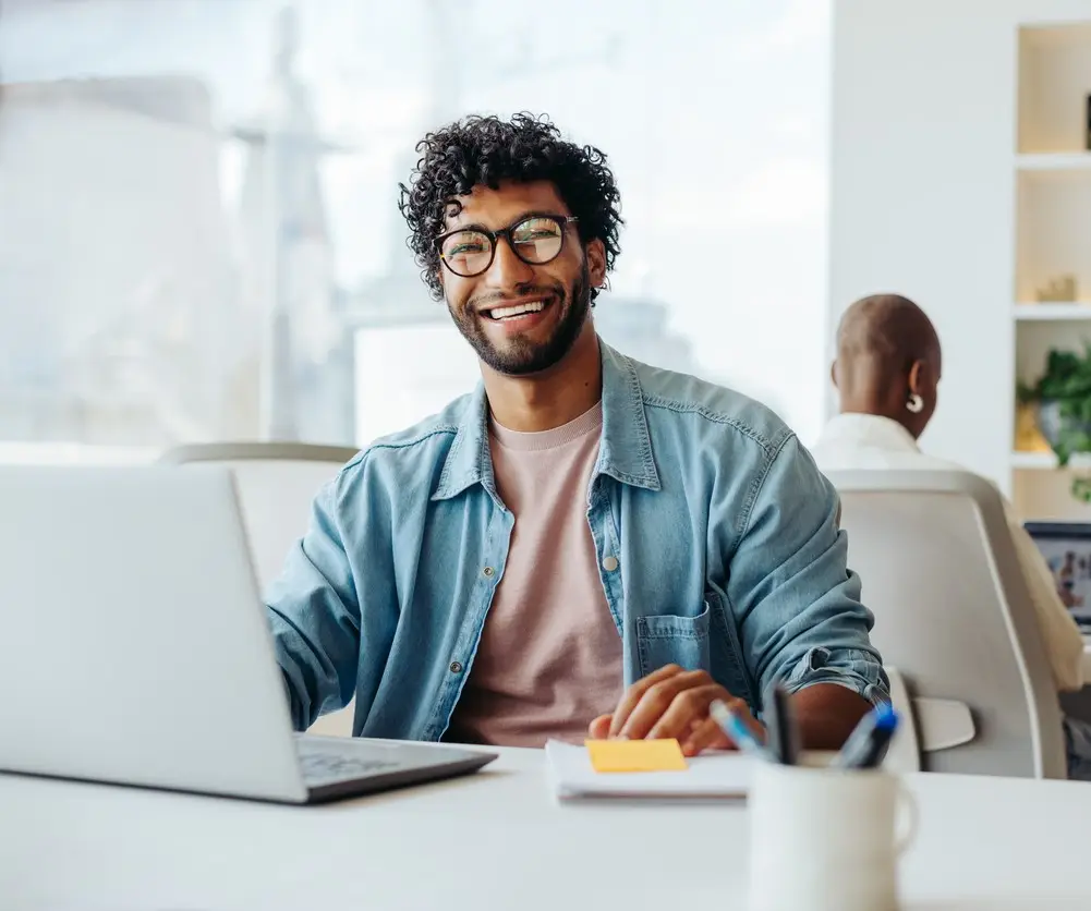 Young male entrepreneur with curly hair and glasses sits at a modern office table. He is working on a laptop and appears happy and smiling. The professional is surrounded by technology and is successful in his startup business.