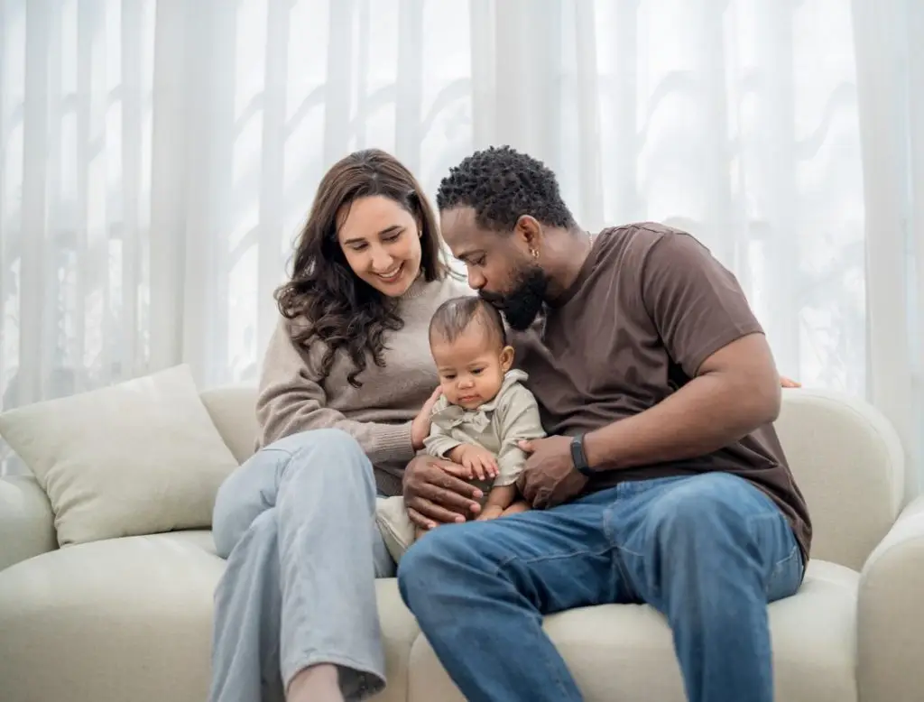 A couple sits closely on a soft sofa, sharing a joyful moment with their baby. The bright room features large windows, creating a warm atmosphere for family bonding.