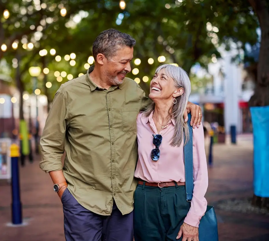 Happy multiethnic man and smiling woman enjoy a romantic evening outdoors. Mature couple shares a loving moment while walking in a european city. Smiling senior couple walks closely together under string lights with copy space.