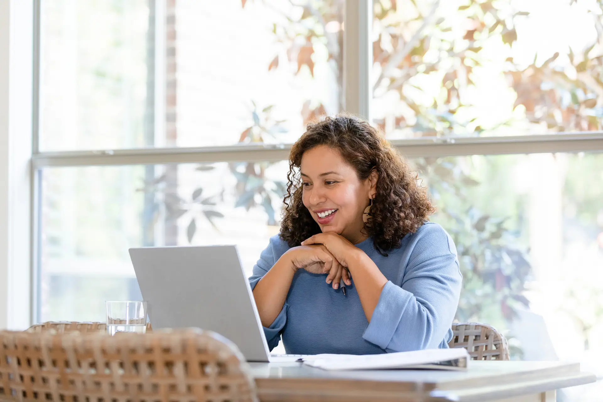 The mid adult woman sits at her dining room table to talk to her family on a video call on the laptop.