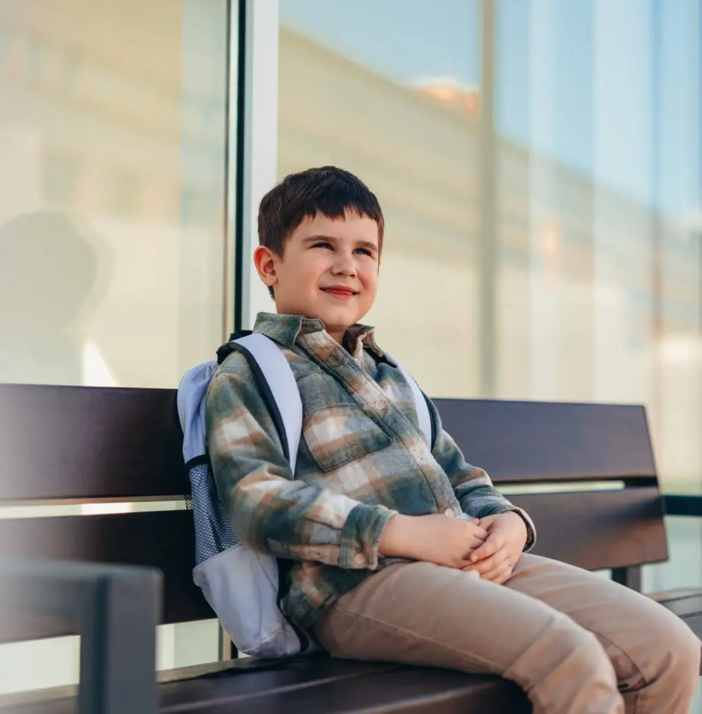 Elementary age boy sitting on a bench outdoors, waiting for school to start, looking away with a smile, embodying the joy and anticipation of childhood in a vibrant city setting