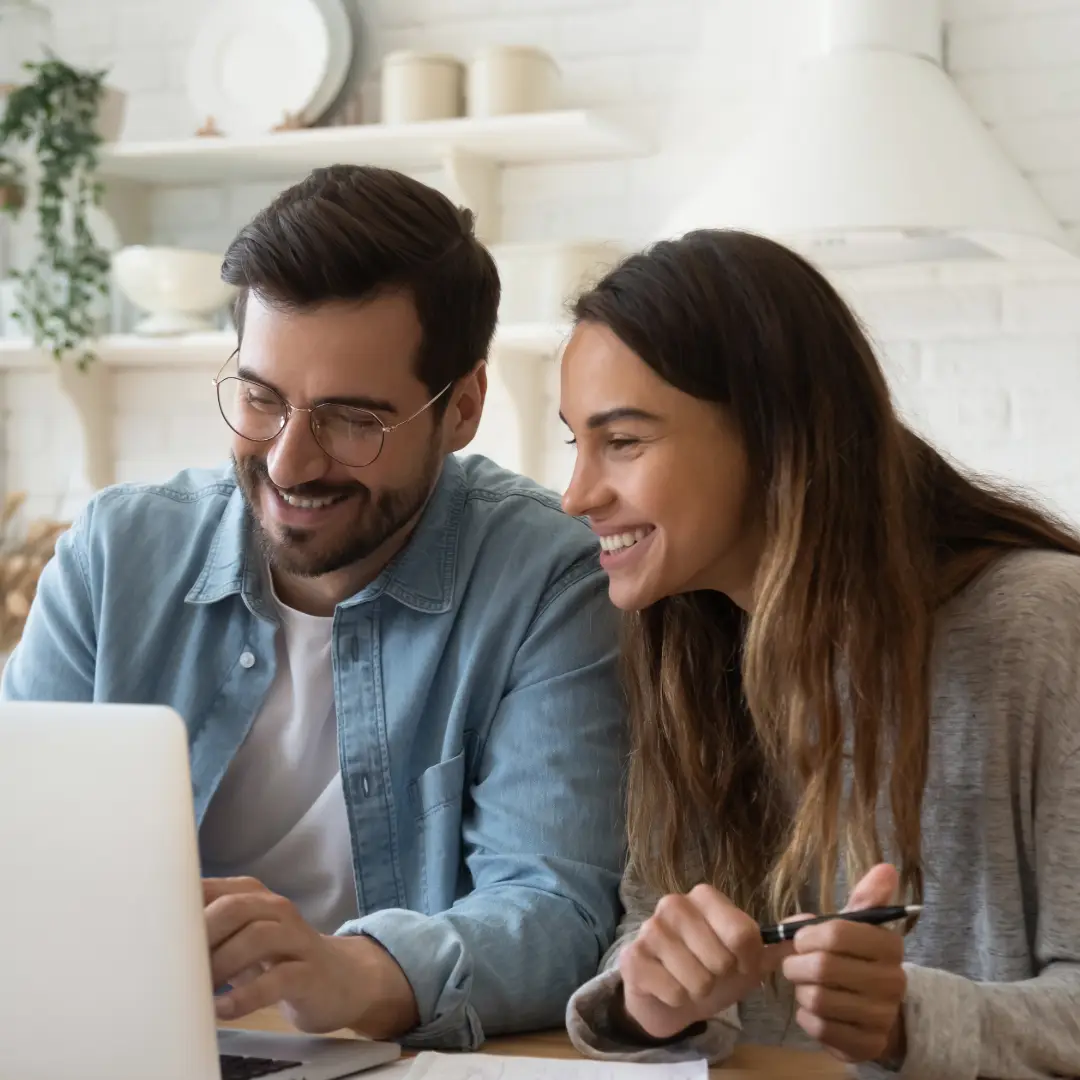 A smiling couple sitting at the kitchen table and looking at a laptop together.