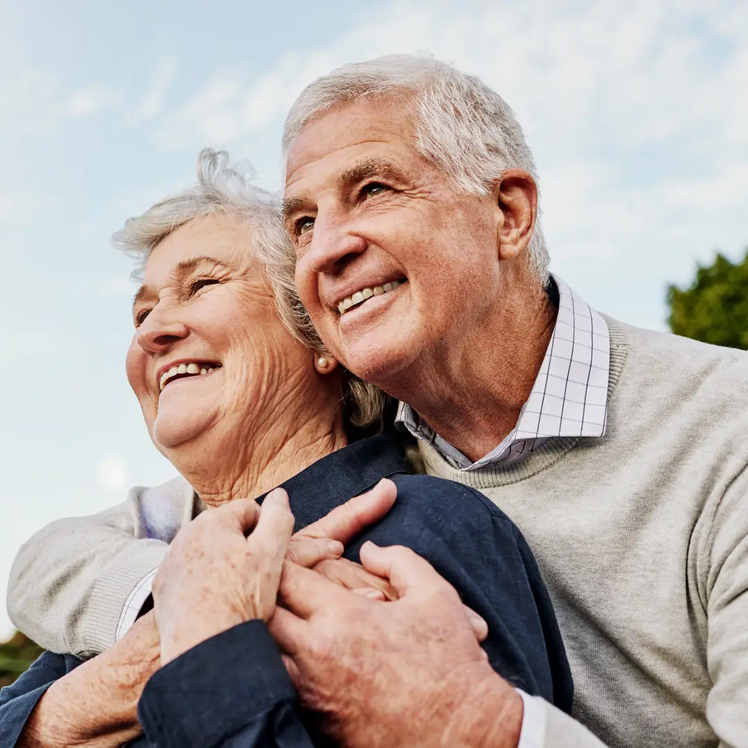 An older couple hugging and smiling with clouds behind them in the sky