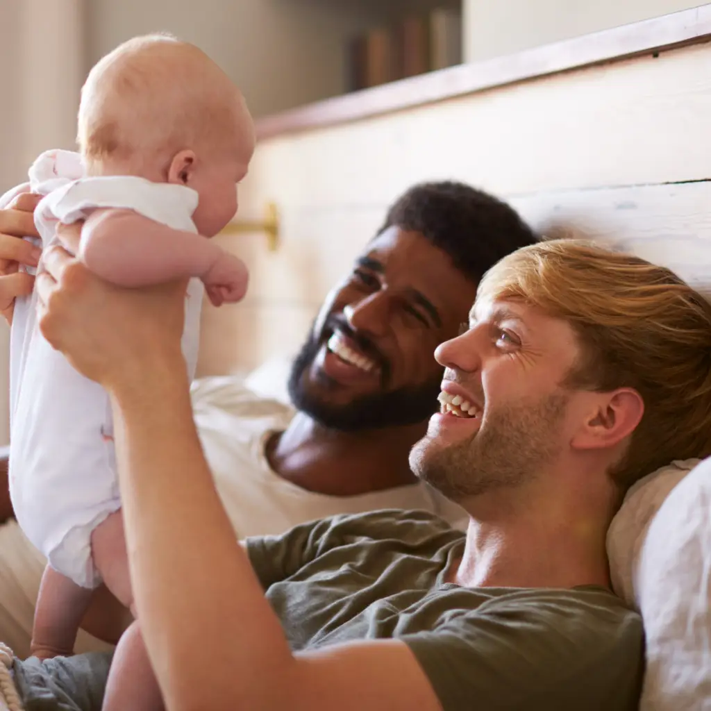 Male couple laughing while laying in bed and playing with their baby