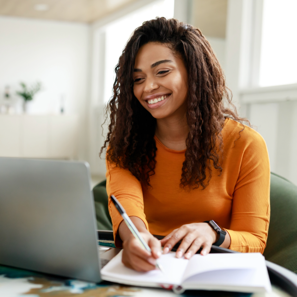 Woman sitting at a kitchen table writing in notebook and looking at a laptop