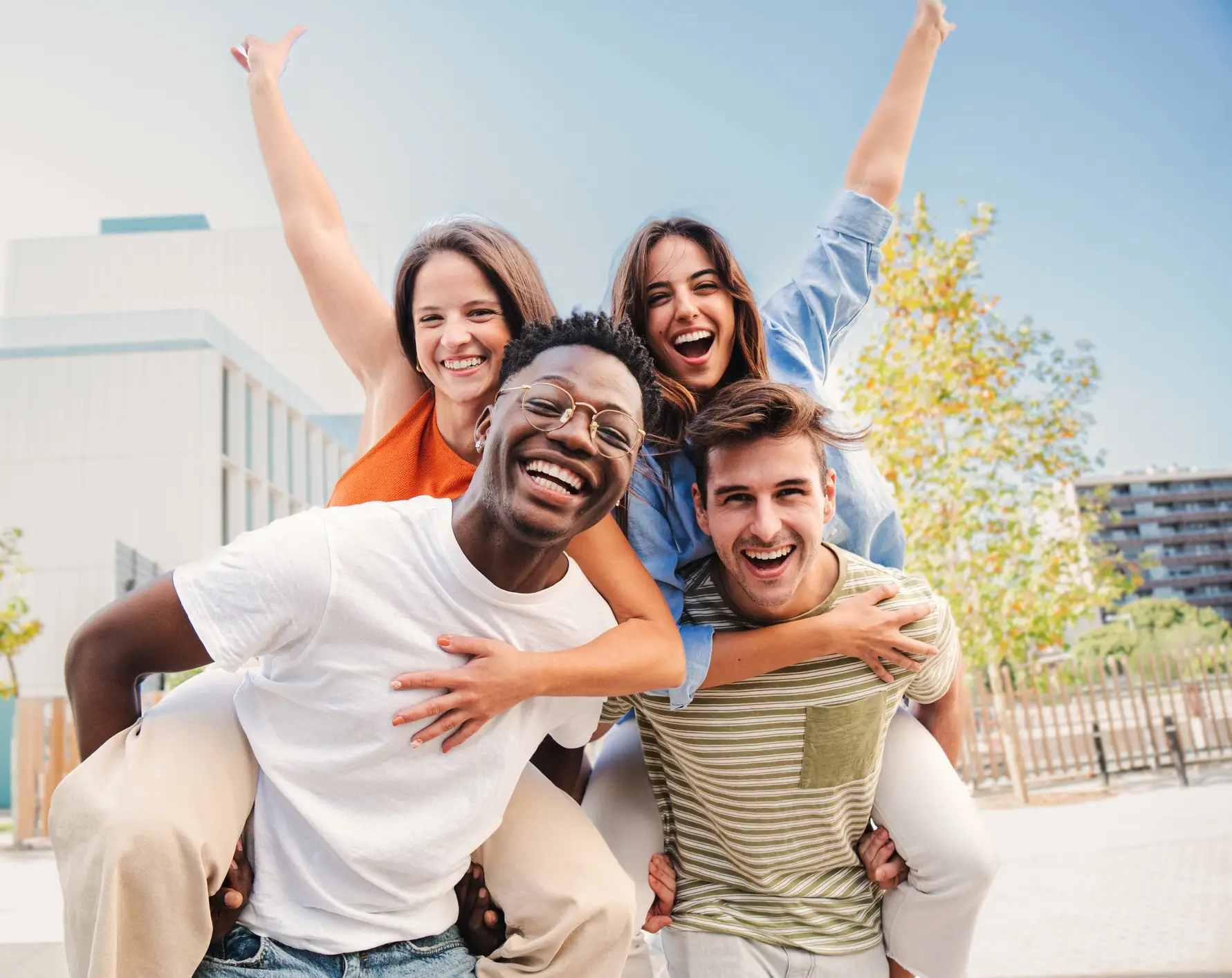 Two multiracial young friends giving piggybacks ride to his girlfriends. A group of happy people having fun and smiling at weekend activity. Men carrying women on his shoulders looking at camera