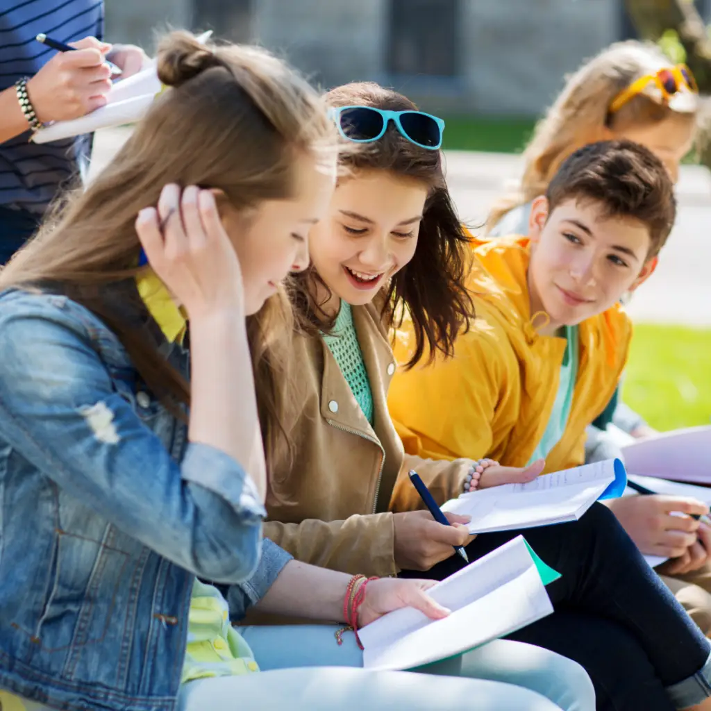 Smiling teens sitting on the grass and doing homework at school