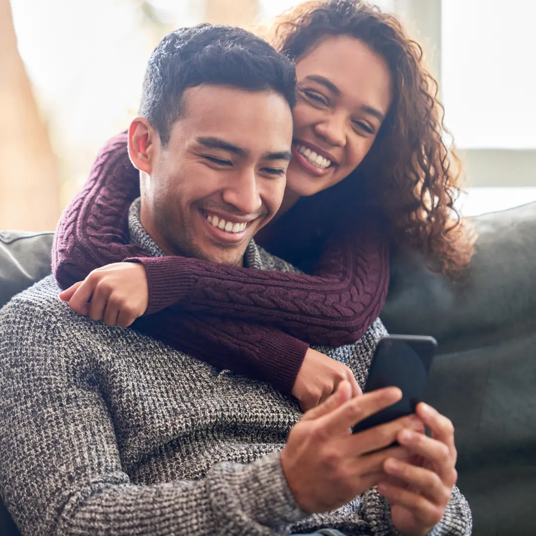 Smiling young woman leaning over the back of the couch and hugging her partner from behind.
